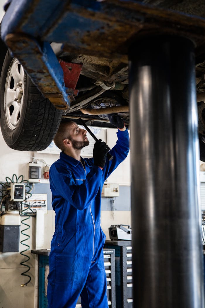 creative Mechanic in blue coveralls inspecting car undercarriage on hydraulic lift.